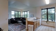 This single-floor space is designed around an open-concept layout that connects the living area and kitchen in one cohesive, flowing environment. The living area is anchored by a large, dark sectional sofa arranged atop a richly patterned area rug, creating a comfortable and inviting atmosphere. A glass-top coffee table adds a modern touch to the seating arrangement. Natural light pours in generously through an array of large, multi-pane windows with black frames, offering views of lush greenery outside and lending the space a bright, airy feel. The vaulted ceiling adds a sense of vertical openness, enhancing the overall spaciousness of the room. A peninsula counter serves as a natural dividing element between the living and kitchen areas. The kitchen features warm wood cabinetry, a double sink, and tile flooring with a classic checkered pattern, offering a functional and well-organized workspace. Light-toned walls and neutral finishes throughout maintain a clean, understated aesthetic. The overall ambiance of the space is warm and welcoming, with a balance of natural materials and modern design elements that create a comfortable, livable environment suitable for both daily routines and relaxed gatherings.