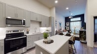 This single-floor residence offers a thoughtfully designed open-concept layout that seamlessly connects the kitchen, dining, and living areas. The kitchen is a standout feature, boasting tall grey shaker-style cabinetry that extends toward the soaring ceilings, stainless steel appliances including a French door refrigerator, electric range, and built-in microwave, all complemented by crisp white quartz countertops and a classic subway tile backsplash. A generously sized kitchen island provides additional prep space and casual seating, making it an ideal hub for entertaining. Wood-look flooring runs throughout the main living areas, adding warmth and continuity to the space. Recessed lighting illuminates the open layout, while large windows in the living area allow natural light to flow in and offer views to the outdoors. A ceiling fan with a pendant-style fixture adds a modern touch overhead. The bedroom features a bold design with deep navy accent walls and plush carpeting, creating a cozy and sophisticated retreat. A large window with blinds and drape panels brings in natural light while maintaining privacy. The bathroom is finished with wood-look flooring, a sleek vanity with white countertops, a glass-enclosed walk-in shower adorned with marble-style tile, and convenient access to a dedicated walk-in closet with built-in shelving and hanging rods.