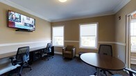 This single-story office space offers a well-appointed and functional environment suited for both individual work and small collaborative gatherings. The room is finished with warm tan walls accented by white crown molding and a chair rail, lending a polished, professional aesthetic. Blue-gray patterned carpeting grounds the space and adds a contemporary contrast to the warmer wall tones.

Two double-hung windows fitted with roller shades allow natural light to filter in while offering privacy and glare control. The views outside suggest a verdant, tree-lined setting. A wall-mounted flat-screen television is positioned near the entrance, making it practical for presentations or virtual meetings.

The layout accommodates multiple functions simultaneously. A dedicated desk workstation with an ergonomic chair and storage unit sits along one wall, while a round conference table with additional seating occupies another area, supporting small group discussions. A comfortable upholstered armchair positioned between the windows provides an informal seating option. Overhead flush-mount lighting ensures consistent illumination throughout. Overall, the space balances functionality with a refined, welcoming atmosphere.