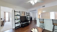 This 2-bedroom, 1-bathroom residence is thoughtfully designed to maximize comfort and functionality across a single floor. The living area serves as the heart of the home, featuring rich hardwood-style flooring, a ceiling fan with light fixture, and a textured ceiling that adds character to the space. Open shelving with ladder-style bookcases provides both storage and display opportunities, lending the room a modern, organized aesthetic.

A seamless flow connects the living area to the dining space, visible through an open archway that enhances the sense of openness throughout. The kitchen is positioned conveniently adjacent to the living room, separated by a breakfast bar with seating — ideal for casual dining or entertaining. Warm lighting from a table lamp creates an inviting ambiance in this area.

The dining room, visible in the background, accommodates a full dining set, making it well-suited for everyday meals or hosting guests. Throughout the unit, the consistent use of dark-toned furniture against light walls creates a clean, contrasting visual appeal. The layout is efficiently arranged to provide a comfortable living experience, with each area flowing naturally into the next.