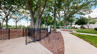 Located at 500 East Constitution Drive in Palatine, Illinois, this well-appointed outdoor recreational space offers a comprehensive array of amenities set within a beautifully landscaped environment. The grounds are framed by a mature canopy of large deciduous trees that provide ample shade and a park-like atmosphere throughout the property.

A full-size basketball court finished in teal and blue surfacing serves as a central recreational feature, surrounded by tall arborvitae hedges and additional mature trees that offer a natural enclosure. Adjacent to the court, a dedicated outdoor fitness area is equipped with multiple green exercise stations installed on rubberized red surfacing, making it suitable for year-round physical activity. Winding concrete pathways connect the various zones of the property, guiding visitors through the well-maintained green lawns and landscaped garden beds.

The building exteriors are finished in white brick and feature clean, modern lines accented with dark trim. Thoughtfully designed garden beds near the building entrances showcase flowering plants and ornamental grasses, adding color and curb appeal. Black wrought iron fencing delineates portions of the grounds, providing structure without obstructing sightlines. The overall ambiance is green, open, and inviting, offering residents a comprehensive outdoor environment for recreation, exercise, and leisure.