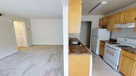 This single-floor space offers a straightforward and functional layout designed for comfortable everyday living. The kitchen is equipped with light wood cabinetry, dark granite-style countertops, a stainless steel sink, and a full-size white refrigerator. A partial wall divides the kitchen from the adjacent living area, creating a sense of separation while maintaining an open feel between the two spaces. The kitchen features tile flooring and overhead fluorescent lighting, providing a bright and practical workspace. A doorway off the kitchen leads to an additional area, suggesting further utility space or storage. The living area is generously sized, finished with wall-to-wall carpeting in a neutral tone that lends a warm and understated ambiance to the room. Neutral-painted walls throughout the space contribute to a clean, move-in-ready aesthetic. The overall design is simple and efficient, with a focus on usability and open flow between the main living and kitchen zones. The single-floor configuration ensures easy accessibility throughout the entire space.