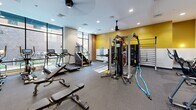 Located at 309 W Front St in Traverse City, Michigan, this fitness center offers a comprehensive workout facility within a single, open floor plan. The space is thoughtfully designed with a bold two-tone color scheme — a vibrant mustard yellow upper wall paired with crisp white below — creating an energetic and motivating atmosphere throughout.

Large floor-to-ceiling windows line one side of the room, flooding the interior with abundant natural light and offering views of the surrounding streetscape. Exposed brick accents add an industrial character that complements the modern finishes and recessed lighting above.

The fitness floor is fully outfitted to accommodate a wide range of training styles. Cardio equipment includes Spirit-brand treadmills, an elliptical, a stair climber, and a stationary bike. Strength training is well-represented with a dual cable functional trainer, a multi-station rack system with suspension training attachments, and a dedicated free weight area featuring a dumbbell rack and adjustable benches. Functional training accessories such as battle ropes, medicine balls, a punching bag, stability balls, and a BOSU balance trainer round out the equipment selection.

A large wall mirror expands the sense of space and aids in form monitoring. Durable rubber flooring covers the entire workout area, and ceiling fans provide additional air circulation. Decorative framed artwork adds a polished finishing touch to this well-appointed fitness facility.