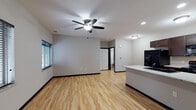 This single-story unit is designed with an open-concept layout that seamlessly connects the living area and kitchen, creating a spacious and functional flow throughout the main living space. Light hardwood flooring extends across the living and kitchen areas, contributing to a bright and clean aesthetic, while neutral gray walls provide a versatile backdrop throughout.

The kitchen is well-equipped, featuring dark espresso cabinetry that offers generous storage, light granite-patterned countertops, and a peninsula with a built-in double sink that doubles as a breakfast bar. Black appliances — including a side-by-side refrigerator with ice and water dispenser, range, microwave, and dishwasher — complement the cabinetry. A door at the rear of the kitchen provides additional access.

The living area is generously proportioned, receiving natural light through horizontal-slatted blinds on multiple windows. A ceiling fan with integrated lighting is centrally positioned overhead, enhancing both comfort and illumination.

The bedroom features plush carpeting, a ceiling fan, and a window with blinds for natural light and privacy. The bathroom is finished with dark wood cabinetry, a granite-topped vanity, a tall linen tower, a large mirror, and a full tub-shower combination, all set on tile flooring.
