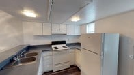 This two-floor space offers a straightforward and functional layout designed for everyday living. The kitchen is a standout feature, showcasing crisp white cabinetry that lines the upper and lower walls, providing generous storage throughout. Dark laminate countertops contrast cleanly against the white finishes, while the space is equipped with a full suite of appliances including a coil-top electric range, a built-in range hood, and a full-size refrigerator. A sink is conveniently positioned along the counter, and the layout follows a practical U-shaped configuration that maximizes workspace efficiency. Track lighting installed along the ceiling, paired with a flush-mount light fixture, ensures the kitchen is well-lit. A small window allows for a degree of natural light to filter into the space. The flooring features dark wood-look planks that ground the room with a warm, contrasting tone against the lighter cabinetry and walls. The textured ceiling adds a subtle architectural detail throughout. Overall, the space presents a clean, utilitarian aesthetic with a neutral palette that provides a versatile foundation across both floors.