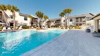 Located at 77 Exuma Way in Santa Rosa Beach, Florida, this outdoor amenity complex offers a well-appointed, resort-style environment designed for relaxation and community living. The centerpiece is a generously sized swimming pool with crystal-clear water, framed by a broad, paver-tiled sun deck that provides ample space for lounging. Aqua-colored chaise lounge chairs and shaded umbrella stations are arranged throughout the deck, complementing the coastal aesthetic. A separate spa pool offers an additional retreat within the same well-landscaped setting.

Mature sabal palms are interspersed throughout the pool area, adding a tropical character while providing natural shade. Neatly maintained landscaping with manicured shrubs, native grasses, and fresh pine straw borders the walkways and fenced perimeters, creating a polished yet inviting atmosphere.

The adjacent single-story clubhouse building features a distinctive whitewashed brick facade accented by dark-framed windows, exposed wood rafter tails, and traditional copper gas lanterns — a nod to coastal Southern architectural style. The surrounding multi-story residential buildings are finished in crisp white with dark window trims and screened balconies, maintaining a cohesive, upscale coastal community aesthetic throughout the property.