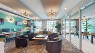 Located at 3015 N Rocky Point Dr E in Tampa, Florida, this single-floor amenity space serves as a sophisticated communal hub designed for both productivity and relaxation. The interior is anchored by a bold contemporary aesthetic, combining teal and charcoal accent walls with warm gold-toned fixtures and layered, patterned carpeting throughout. Statement sputnik and sculptural chandelier lighting illuminate each zone, adding a touch of upscale flair. The main lounge area features an arrangement of plush dark leather club chairs and a velvet sofa grouped around a central coffee table, with floor-to-ceiling windows that invite abundant natural light and views of the surrounding Tampa landscape, including palm trees and neighboring waterfront buildings. A dedicated business center nook lines one wall with built-in desks, computer workstations, and printer access, illuminated by artistic wall sconces and framed by large windows. Adjacent to the main lounge, a private conference or meeting room offers dark dining-style tables paired with bold animal-print accent chairs, a mounted flat-screen TV, and richly textured teal wallcovering with coordinating gold-loop chandelier lighting. Glass partition walls connect the zones while maintaining visual openness, and lush potted tropical plants reinforce the space's vibrant, resort-inspired ambiance.