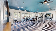 Located in Wildwood, FL, this fitness center is a thoughtfully designed exercise facility that combines functionality with striking aesthetics. The space is immediately distinguished by its bold teal-painted ceiling, recessed lighting, and multiple ceiling fans that ensure comfortable airflow throughout. A vibrant geometric patchwork carpet in shades of blue, gray, and beige covers the floor, adding a dynamic visual element to the room.

Large floor-to-ceiling windows and glass doors line one wall, flooding the space with natural light and offering views of the surrounding outdoor area. Full-length mirrors, framed by elegant arched accents, are strategically placed to visually expand the room and support proper form during workouts.

The exercise floor is comprehensively outfitted with a wide range of equipment, including treadmills, ellipticals, stationary bikes, a cable machine, multi-use strength machines, free weights, a weight bench, and stability balls. Decorative laser-cut panels provide a stylish partition between equipment zones.

An accent wall features an arched niche with an illuminated motivational sign, a mounted flat-screen TV, and a beverage refrigerator, creating an engaging focal point. A water station is also available for convenience. The overall ambiance is energetic yet polished, making this a well-rounded fitness destination.