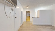 Located at 5119 Fountain Ave in the heart of Los Angeles, CA, this efficiently designed single-floor unit offers a thoughtful layout comprising a living room, kitchen, and bathroom. Light wood laminate flooring runs consistently throughout, lending warmth and visual continuity to the space.

The living area is bright and open, featuring white walls, a textured ceiling, a wall-mounted AC unit, and a large window with horizontal blinds that allows natural light to fill the room. The entry flows naturally into the rest of the unit, aided by a clean, minimal design aesthetic.

The kitchen is well-equipped with white cabinetry, dark granite-style countertops, a stainless steel gas range, a ventilation hood, and a striking dark subway tile backsplash. A window above the counter area brings in additional natural light and views of surrounding greenery. A peninsula counter separates the kitchen from the main living space, adding functional division without enclosing the area.

The bathroom is split into two areas: a vanity section with a floating white cabinet, dark countertop, dual mirrors, and a single sink; and a separate wet area with a soaking tub and shower combination, toilet, and a frosted window for privacy. The consistent light flooring ties both areas together seamlessly.

Overall, the space presents a modern, clean, and functional environment suited for comfortable urban living in Los Angeles.