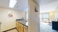 Located at 1705 SW 11th Ave in Portland, Oregon, this one-bedroom, one-bathroom unit offers a well-organized layout across a single floor. The living room serves as the heart of the unit, featuring warm yellow-toned walls, patterned carpet tile flooring, and a large sliding glass door that floods the space with natural light and provides views of the surrounding greenery. The open-concept kitchen connects seamlessly to the living area, equipped with light wood cabinetry, a breakfast bar with seating, and standard appliances including a refrigerator and stove — a practical setup for everyday living. The bedroom is a comfortable, well-proportioned room accommodating two twin beds, each paired with a dedicated desk and task lighting, making it a functional space for study or work. A full-panel wood closet provides ample storage, and a window allows for additional natural light. The bathroom features light wood cabinetry, an integrated sink and vanity, and a shower/tub combination, finished in warm, neutral tones consistent throughout the unit. A hallway connects the main spaces efficiently. The unit's cohesive warm yellow palette and carpet tile flooring create a consistent, welcoming ambiance throughout.