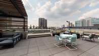 This expansive rooftop terrace offers a well-appointed urban outdoor retreat designed for both relaxation and social gatherings. The space is thoughtfully organized into several distinct zones, each serving a unique purpose while contributing to the overall cohesive aesthetic.

At the heart of the terrace, a striking pergola structure with slatted wood panels and a dark steel frame shelters a built-in bar with warm wood cladding and a sleek dark countertop. Adjacent to the bar, an outdoor kitchen equipped with a grill provides ample space for entertaining. Beneath the pergola, a sophisticated dark lounge set with plush cushions creates an inviting conversation area.

A raised section of the terrace features built-in bench seating accented with colorful throw pillows in teal, yellow, and patterned fabrics, alongside a fire pit surrounded by patterned lounge chairs. Aqua-toned bistro tables and chairs are scattered throughout, adding a playful pop of color against the gray tile flooring.

The terrace is enclosed by sleek metal railings and bordered by planter boxes filled with flowering greenery. Mounted televisions and modern wall-mounted lighting complete the amenity-rich environment. Throughout the space, panoramic views of the surrounding urban skyline provide a dramatic backdrop, making this rooftop terrace a premier destination for outdoor living.