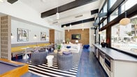 Located at 500 West Camelback Road in Phoenix, Arizona, this single-floor communal lounge is a thoughtfully designed social space that blends modern aesthetics with vibrant, playful touches. The open-plan layout is anchored by soaring ceilings adorned with bold dark wood exposed beams, which contrast dramatically against bright white walls and create an airy, loft-like atmosphere. Striking blue terracotta floor tiles run throughout, adding a warm Southwest-inspired character underfoot.

The living area features plush, neutral-toned sectional seating arranged atop a bold black-and-white striped area rug, centered around a textured accent wall with a mounted flat-screen television. A suspended egg chair and layered decorative accessories contribute to the eclectic, resort-style ambiance. Adjacent to the lounge, a full-size pool table invites recreation, while a built-in wet bar with under-counter appliances provides convenient amenity access.

A dining nook with yellow upholstered banquette seating and bistro-style tables offers a casual gathering spot. Floor-to-ceiling black-framed windows and glass doors flood the space with natural light and frame views of the outdoor pool area and landscaped surroundings. Curated artwork and globe pendant lighting complete the sophisticated yet approachable design of this exceptional communal retreat.