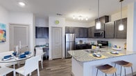 This single-floor residence showcases a thoughtfully designed open-concept layout that seamlessly connects the kitchen, dining, and living areas. The kitchen is a standout feature, equipped with stainless steel appliances including a refrigerator, microwave, and range. Rich, dark cabinetry provides ample storage, while granite countertops add an elegant touch. A peninsula with seating extends the counter space and serves as a natural divider between the kitchen and dining area, making it ideal for casual meals or entertaining.

Pendant lighting and a track light fixture illuminate the kitchen, enhancing the overall brightness of the space. The flooring throughout is a warm-toned wood-style material that adds continuity and a contemporary feel across the open areas. The dining area accommodates a table and chairs, positioned near a built-in desk nook that offers a practical workspace. White walls provide a clean, neutral backdrop that contrasts nicely with the darker cabinetry and flooring. The overall ambiance is modern and functional, with a cohesive design aesthetic throughout.