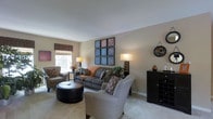 This single-floor space showcases a well-appointed living room that serves as the heart of the home. The room is designed with a neutral color palette of warm beige and taupe tones, creating a calm and welcoming ambiance throughout. Plush, patterned seating — including a sofa, accent chairs, and a tufted leather ottoman — are arranged to encourage comfortable conversation and relaxation. Natural light filters in through a window dressed with a classic Roman shade, brightening the carpeted space. The walls are thoughtfully decorated with a bold grid of framed artwork as a central focal point, complemented by a round decorative mirror and additional wall accents. A pair of matching table lamps provides warm ambient lighting, enhancing the cozy atmosphere during evening hours. A dark wood storage cabinet adds both function and contrast against the lighter walls, while potted greenery introduces a natural, refreshing element to the room. The overall design balances decorative detail with practical comfort, making this a versatile and stylish living area suited for both everyday use and entertaining guests.
