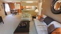 This single-floor residence showcases an open-concept design that seamlessly connects the living, dining, and kitchen areas into one cohesive space. The living room is carpeted and comfortably sized, anchored by a tufted sofa and a cushioned armchair arranged around a dark coffee table, creating an inviting lounge area. A warm accent wall in a rich terracotta orange adds a bold visual focal point, complementing the neutral tones of the furnishings and carpet throughout.

A pass-through counter connects the living area to the kitchen, which features white cabinetry, a stainless refrigerator, and a range, offering an efficient and accessible layout. The open sightlines between the kitchen and living space make the area feel expansive and well-suited for everyday living and entertaining.

Adjacent to the living room, the dining area accommodates a round table with seating for several guests, enhanced by the same warm color palette that unifies the space. Recessed and overhead lighting throughout ensures the interior remains bright and well-lit. Overall, the space presents a practical and cohesive design with warm tones and an open, connected floor plan.
