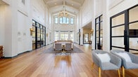 This stunning communal space makes an immediate impression with its dramatic vaulted, cathedral-style ceiling accented by exposed beams and a grand gold chandelier that anchors the main gathering area. The open, single-floor layout is expansive, offering generous room for a variety of seating arrangements and social zones. Wide-plank hardwood flooring flows throughout, lending warmth and continuity to the bright, white interior.

Floor-to-ceiling black-framed glass partition walls divide dedicated areas — including a private office or conference room visible through the glazing — while maintaining a sense of openness and visual connectivity. Tall, multi-pane windows line the perimeter, flooding the interior with abundant natural light and offering views of the surrounding landscape.

The main lounge area features a curated mix of contemporary furnishings, including plush sofas, tufted armchairs in neutral tones, and gold-accented accent tables. A separate game and recreation zone houses a shuffleboard table, while cozy lounge nooks with built-in banquette seating offer intimate gathering spots. A textured wood console near the entry, paired with large-scale artwork, adds a distinctive artistic touch.

Wall sconces with gold fixtures, recessed lighting, and thoughtfully placed décor complete the refined, modern-transitional aesthetic of this welcoming and highly functional community space.