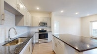 This single-story home offers a thoughtfully designed layout across a comfortable footprint, accommodating three bedrooms, one full bathroom, and two closets connected by a central hallway. The kitchen serves as a functional centerpiece, featuring white shaker-style cabinetry with brushed gold hardware, dark granite countertops, a classic white subway tile backsplash, and a full suite of stainless steel appliances including a French door refrigerator, electric range with over-the-range microwave, and dishwasher. A kitchen island provides additional counter and storage space, while light hardwood-style flooring extends throughout the kitchen and living areas, enhancing the bright, clean aesthetic. Recessed lighting illuminates the space effectively throughout. The living area benefits from natural light through multiple windows and includes direct exterior access via a door. The bedrooms are carpeted for comfort, with the generously sized primary bedroom offering ample space. Secondary bedrooms include ceiling fans and windows with outdoor views. The full bathroom is finished with a white shaker vanity, dark granite countertop, tub-shower combination, and built-in open shelving for added storage convenience.