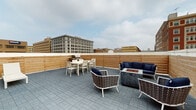 This rooftop terrace offers a well-appointed urban outdoor retreat set atop a building surrounded by a classic cityscape. The floor is finished with gray paver tiles throughout, providing a clean, durable surface that anchors the space. A horizontal wood-slatted perimeter fence runs along the edges, offering both privacy and a warm, natural contrast to the industrial urban backdrop visible beyond its borders—including multi-story historic brick and masonry buildings.

The terrace is organized into distinct functional zones. A lounge area is centered around a rectangular gas fire pit table, furnished with woven rattan-style chairs and a navy blue cushioned sectional sofa, creating an inviting setting for relaxed gatherings. A separate dining zone features a white table with matching chairs and a gas grill station, ideal for outdoor cooking and al fresco meals. Additional seating, including white chaise lounges, is positioned to take advantage of the open sky and surrounding views.

The building's bold red brick exterior wall frames one side of the terrace, adding architectural character to the space. Overall, the terrace presents a functional and stylish outdoor environment well-suited for both casual lounging and social entertaining in an urban setting.