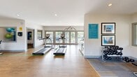 Located at 936 Mountain Creek Road in Chattanooga, Tennessee, this well-appointed fitness center at Ascent at Signal Mountain offers a thoughtfully designed single-floor layout with generous open space for a full range of workout activities. The main exercise area features wood-look flooring with bold yellow lane markings, providing a clean and energetic aesthetic throughout. Cardio equipment, including treadmills, elliptical machines, and a stair climber, is arranged to make efficient use of the space, while a dedicated strength area with free weights and a weight bench sits atop a durable carpet tile zone. A large wall-mounted mirror enhances the sense of depth and supports strength training. Multiple flat-screen TVs are positioned throughout the room, including on a vibrant teal accent wall, keeping members entertained during workouts. Natural light pours in through large French doors and windows that open onto an outdoor patio, offering pleasant views of the surrounding greenery. A stylish wet bar station, accented by striking teal subway tile, includes a sink and small refrigerator, providing convenient hydration access. Recessed lighting and crisp white walls complete the modern, motivating ambiance of this community fitness facility.