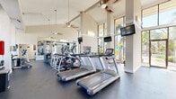 Located at 601 Rosery Rd NE in Largo, Florida, this fitness center offers a generously proportioned, single-floor exercise facility designed to support a comprehensive workout experience. The main exercise room is defined by its dramatically vaulted ceilings with exposed beams, ceiling fans, and an abundance of natural light streaming through floor-to-ceiling windows and glass doors. These expansive windows frame views of the surrounding palm trees and outdoor areas, creating an energizing, resort-like atmosphere throughout.

The exercise room is well-equipped with a range of cardio machines, including multiple treadmills and elliptical trainers, as well as a cable resistance machine suitable for full-body strength training. Wall-mounted televisions are positioned throughout for entertainment during workouts. Mirrored walls enhance the sense of space and provide functional feedback for exercise form. The dark rubberized flooring adds both durability and a polished, professional aesthetic.

A vibrant large-scale painting serves as a bold focal point on one wall, adding an artistic touch to the otherwise clean, neutral interior. A connecting hallway provides access between spaces. The overall design balances functionality with an uplifting, light-filled ambiance, making this fitness center a well-appointed and inviting space for residents and users alike.