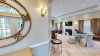 The Fairfield Townhouses at Bohemia community clubhouse, located at 100 Saddle Cove Road in Bohemia, NY, offers residents a refined and well-appointed gathering space spread across a single floor. The entrance foyer sets an elegant tone with warm sconce lighting, an ornate gold-framed mirror, and classic wainscoting that carries throughout the space.

The main lounge area is a standout feature, showcasing a white-mantled fireplace with a rich stone surround, a wall-mounted flat-screen television, and a tray ceiling with recessed lighting. Soft sage-green walls paired with neutral tile flooring create a calm, inviting atmosphere. Plaid armchairs, upholstered seating, and glass-topped accent tables furnish the room comfortably for socializing or relaxing.

A separate seating area offers a beautiful bay window with arched transoms, floor-to-ceiling draperies, and additional casual seating, while large French doors with arched windows open to an outdoor pool area, flooding the interior with natural light.

The fitness center is fully equipped with multiple treadmills, elliptical machines, and stationary bikes, all set on durable red carpet. Wall-to-wall mirrors, flat-screen monitors, elevated ceilings, and arched windows with natural light make this an energizing workout environment. Together, these amenity spaces reflect a polished, community-focused design.