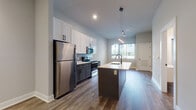 This contemporary single-floor residence is thoughtfully designed with a clean, modern aesthetic throughout. The open-concept kitchen serves as a central highlight, featuring two-tone cabinetry with white upper shaker-style cabinets and dark lower cabinets, stainless steel appliances including a refrigerator, electric range, and over-the-range microwave, and a large central island with a light countertop and integrated sink. Pendant lighting over the island adds a stylish focal point, while hardwood-style flooring flows seamlessly throughout the main living areas, enhancing the sense of continuity and warmth. The living area benefits from soaring ceilings that lend an airy, expansive feel, with natural light streaming in through large windows that offer views of the surrounding greenery. Bedrooms feature plush carpeting and generously sized windows with blinds, creating comfortable and well-lit retreats. The bathroom is finished with modern fixtures, including a vanity with a white countertop, black hardware accents, and wood-look flooring that ties the space together. Additional highlights include a dedicated laundry area and ample closet storage, making this a highly functional and stylish residence throughout.