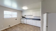 This single-floor, 3-bedroom, 2-bathroom garden unit offers a straightforward and functional layout with clean, neutral finishes throughout. The kitchen is well-equipped with ample white cabinetry, dark countertops, a white electric range, refrigerator, and a double-basin stainless steel sink. A white subway tile backsplash adds a classic touch, while wood-look flooring grounds the space with a warm, durable surface. Natural light filters in through a window fitted with blinds, brightening the cooking area. The open adjacent area provides generous room for dining or additional living space, also finished with the same wood-look flooring. The spacious living area features plush carpet and a large window that allows for plenty of natural light, creating a comfortable and welcoming environment. Each of the three bedrooms is carpeted and painted in soft gray tones, offering a calm and versatile space. At least one bedroom includes a closet with a hanging rod and direct access to a bathroom. Both bathrooms are finished with white tile surrounds, tub-shower combinations, and textured walls accented with a subtle blue tile border detail. The unit's consistent neutral palette and practical layout make it a functional and comfortable living space.