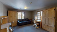 Located at 2205 College Ave in Berkeley, CA, this single-floor unit offers a functional and practical layout suited for residential living near the UC Berkeley campus. The living area is carpeted and furnished with a dark leather sofa, light wood armoires, a built-in desk with chair, and a radiator heater beneath leaded glass windows that allow natural light to filter in. The textured ceiling and neutral gray-toned walls give the space a understated, utilitarian character consistent with collegiate housing.

The kitchen features light wood cabinetry extending to the upper walls, a white electric range, black refrigerator, microwave, and a sink with ample counter space. Dark hardwood-style flooring in the kitchen contrasts with the carpeted living and sleeping areas, clearly delineating the functional zones. The bedroom accommodates two twin beds with wooden frames, positioned on either side of a large multi-pane window that frames outdoor views and fills the room with natural light. A built-in shelf niche adds convenient storage.

The bathroom includes tile flooring and a distinctive diamond-patterned leaded glass window, adding a traditional architectural touch. Overall, the unit presents a straightforward, well-organized living environment with consistent wood accents throughout.