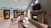 This single-story space is defined by its striking architectural character, anchored by soaring vaulted ceilings that lend an open, airy feel throughout. The main gathering area centers around a handsome stone fireplace with a broad hearth and wooden mantel, complemented by a wall-mounted television above. Rich, variegated hardwood flooring runs throughout, adding warmth and texture to the neutral gray walls.

An abundance of natural light flows in through large, wood-framed windows, including clerestory windows positioned high along the vaulted roofline, offering views of surrounding greenery. A cozy lounge nook near the window wall features a pair of gray leather accent chairs arranged around a round side table, creating an inviting retreat.

The fully equipped kitchen showcases rich dark cabinetry, granite countertops, a decorative mosaic tile backsplash, stainless steel appliances, and a peninsula with bar seating — ideal for casual dining or entertaining. Track lighting and recessed fixtures provide ample illumination.

A separate game room, visible through wood-framed glass doors, houses a billiard table and adds a recreational dimension to the space. Overall, the design balances comfort and sophistication throughout.