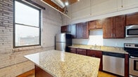 Located at 206 Cesar E Chavez Ave SW in Grand Rapids, Michigan, this single-floor urban loft unit showcases an industrial-chic aesthetic that blends raw architectural character with modern finishes. The open-concept living and kitchen area is anchored by exposed painted brick walls, soaring ceilings with visible timber beams, and industrial-style ductwork, creating a distinctive warehouse-inspired atmosphere. Large windows allow generous natural light to fill the space while framing views of the Grand Rapids skyline.

The kitchen is a standout feature, equipped with rich dark-wood cabinetry, gleaming granite countertops, a substantial center island ideal for dining or food preparation, and a full suite of stainless steel appliances including a refrigerator, range, microwave, and dishwasher. Hardwood flooring extends throughout the main living areas, adding warmth that contrasts beautifully with the industrial elements overhead.

The bedroom offers a quieter retreat with hardwood floors, dark-painted walls, and the same exposed beam ceiling detail seen throughout the unit. The bathroom is finished with consistent granite countertops, dark wood cabinetry, a vanity mirror, and tile flooring, providing a clean and functional space. Overall, the unit delivers a cohesive urban loft experience with upscale finishes throughout.