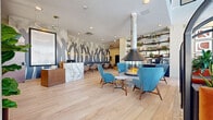 This contemporary single-floor communal space offers a generously proportioned and thoughtfully designed interior that blends modern aesthetics with warm, inviting details. The open-plan layout encompasses a reception area, lounge, dining area, kitchen, and informal meeting spaces, all unified by wide-plank light wood flooring that flows seamlessly throughout.

The centerpiece of the space is a dramatic suspended, 360-degree glass-enclosed fireplace with a cylindrical metallic flue that draws the eye upward toward the bright, recessed-lit ceiling. Surrounding the fireplace, a curated arrangement of mid-century modern lounge chairs in bold teal upholstery with warm wood frames creates an inviting gathering area. 

A statement marble reception desk anchors one side of the space, complemented by an eye-catching abstract wall mural rendered in shades of charcoal and grey. On the opposite end, a continuous banquette finished in deep navy velvet lines the wall, paired with marble-topped bistro tables and globe sconce lighting. Floor-to-ceiling windows flood the space with natural light and provide views of the surrounding neighborhood. Open shelving displays curated décor and greenery, adding warmth and texture. A private nook with a round wood table and leather chairs offers a quiet retreat within the larger space.
