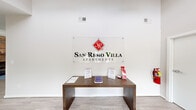 The San Remo Villa Apartments leasing office is a thoughtfully designed single-floor space that balances professional function with a welcoming, residential warmth. Upon entry, visitors are greeted by a bright, open reception area with light hardwood-style flooring and crisp white walls that create a clean, modern atmosphere throughout.

The main gathering area is expansive and airy, anchored by soaring vaulted ceilings and ceiling fans that enhance the sense of openness. A striking floor-to-ceiling brick fireplace with a rustic wood mantel serves as the focal point of the lounge area, where rich brown leather seating is arranged around a blue area rug, lending a comfortable and inviting ambiance.

Adjacent to the lounge, a dedicated office workspace features ample natural light streaming through wide windows with views of the lush landscaping outside. This area is equipped with a large desk and professional seating, providing a functional environment for leasing consultations.

At the rear of the space, French doors open directly to views of the community pool, bathing the interior in abundant natural light and creating a seamless connection to the outdoors. The overall design blends approachable comfort with polished professionalism, making it an ideal setting for prospective residents to explore their new home at San Remo Villa.