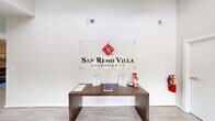 The San Remo Villa Apartments leasing office is a thoughtfully designed single-floor space that balances professional function with a welcoming, residential warmth. Upon entry, visitors are greeted by a bright, open reception area with light hardwood-style flooring and crisp white walls that create a clean, modern atmosphere throughout.

The main gathering area is expansive and airy, anchored by soaring vaulted ceilings and ceiling fans that enhance the sense of openness. A striking floor-to-ceiling brick fireplace with a rustic wood mantel serves as the focal point of the lounge area, where rich brown leather seating is arranged around a blue area rug, lending a comfortable and inviting ambiance.

Adjacent to the lounge, a dedicated office workspace features ample natural light streaming through wide windows with views of the lush landscaping outside. This area is equipped with a large desk and professional seating, providing a functional environment for leasing consultations.

At the rear of the space, French doors open directly to views of the community pool, bathing the interior in abundant natural light and creating a seamless connection to the outdoors. The overall design blends approachable comfort with polished professionalism, making it an ideal setting for prospective residents to explore their new home at San Remo Villa.