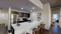 This contemporary single-floor residence showcases a thoughtfully designed open-concept layout that emphasizes clean lines and modern finishes throughout. The kitchen serves as a central highlight, featuring bold dark cabinetry that creates a striking contrast against crisp white quartz countertops. Stainless steel appliances, including a full-size refrigerator, gas range, and built-in microwave, offer both style and functionality. Recessed lighting illuminates the space efficiently, enhancing the sleek aesthetic of the lowered ceiling detail above the kitchen area. A generously sized kitchen island with an integrated sink doubles as a breakfast bar, comfortably accommodating several wooden bar stools for casual dining or entertaining. The warm-toned hardwood flooring grounds the space and adds a natural element that balances the contemporary design palette. Wall-mounted shelving provides a curated display area for artwork and décor, adding personality and warmth to the otherwise clean, modern environment. The open design allows for excellent flow between the kitchen and living areas, creating an inviting and practical living space suited to both everyday living and hosting guests.