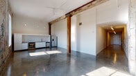 This single-floor industrial loft conversion masterfully balances raw historic character with modern finishes. A striking entry hallway sets the tone immediately, featuring an expansive original brick wall adorned with layers of aged graffiti art, illuminated by overhead track lighting and grounded by gleaming polished concrete floors that extend throughout the space.

The open-plan living and kitchen area is anchored by original heavy timber columns and beams, lending an authentic industrial warmth to the otherwise bright, white-walled interior. The kitchen is fitted with white cabinetry, marble-look countertops, a stainless steel undermount sink with a coil-spring faucet, stainless steel appliances including a dishwasher and refrigerator, and open shelving. Pendant lighting and a ceiling fan complete the functional yet stylish aesthetic.

A dedicated laundry area houses a full washer and dryer alongside the mechanical systems. The two bedrooms offer quiet retreats, one featuring patterned carpeting and an exposed timber post. The two bathrooms are finished in crisp white tile—one with a soaking tub and shower combination, the other with an open walk-in shower, wall-mounted sink, and vanity mirror. Large windows along the original brick exterior wall flood the space with natural light and offer outward views.
