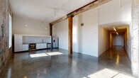 This single-floor industrial loft conversion masterfully balances raw historic character with modern finishes. A striking entry hallway sets the tone immediately, featuring an expansive original brick wall adorned with layers of aged graffiti art, illuminated by overhead track lighting and grounded by gleaming polished concrete floors that extend throughout the space.

The open-plan living and kitchen area is anchored by original heavy timber columns and beams, lending an authentic industrial warmth to the otherwise bright, white-walled interior. The kitchen is fitted with white cabinetry, marble-look countertops, a stainless steel undermount sink with a coil-spring faucet, stainless steel appliances including a dishwasher and refrigerator, and open shelving. Pendant lighting and a ceiling fan complete the functional yet stylish aesthetic.

A dedicated laundry area houses a full washer and dryer alongside the mechanical systems. The two bedrooms offer quiet retreats, one featuring patterned carpeting and an exposed timber post. The two bathrooms are finished in crisp white tile—one with a soaking tub and shower combination, the other with an open walk-in shower, wall-mounted sink, and vanity mirror. Large windows along the original brick exterior wall flood the space with natural light and offer outward views.