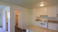 This single-floor residence offers a clean and functional layout designed for comfortable everyday living. The kitchen is a central feature of the space, outfitted with white cabinetry that extends from counter height to the ceiling, providing generous storage. A white electric range and matching appliances give the kitchen a cohesive, uniform appearance. The light-toned countertops offer ample workspace, and a peninsula-style counter extends outward, creating a natural division between the kitchen and the adjoining area while also serving as an informal dining or prep surface. Overhead, a flush-mount ceiling light provides practical illumination throughout the kitchen. The flooring transitions from a lighter surface in the kitchen to rich dark hardwood in the connecting areas, adding warmth and visual contrast to the interior. A doorway visible from the kitchen leads into a well-lit room that appears to include a walk-in closet, suggesting a bedroom with thoughtful storage solutions. The overall palette of the space is neutral and bright, with white walls and ceilings that enhance the sense of openness. The straightforward, efficient design of this residence makes it a practical and inviting place to call home.