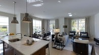 Located at 1-3 Mill River Lane in Ardsley, NY, this single-floor residence offers a thoughtfully designed open-concept living space that seamlessly integrates the kitchen, dining, and living areas into one cohesive environment. The kitchen features a generous island with a light-toned stone countertop and modern pendant lighting, making it a functional centerpiece that anchors the social areas of the home. The dining area accommodates a full table and chairs, positioned to take advantage of the large windows that line the perimeter of the space. The living area is bright and inviting, furnished with a comfortable sofa and complementary seating, all bathed in natural light that streams through multiple large windows and glass doors. Light hardwood flooring flows consistently throughout, reinforcing the open and airy atmosphere. The neutral color palette — featuring soft whites and warm grays — lends the space a clean, contemporary aesthetic. Recessed ceiling lighting supplements the natural light, ensuring the interior remains well-lit at all hours. Views of lush greenery are visible through the windows, connecting the interior to the outdoors and enhancing the overall sense of calm and spaciousness.
