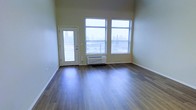This single-floor space presents a clean, modern aesthetic with thoughtful design elements throughout. The main living area is immediately welcoming, featuring rich, dark hardwood-style flooring that contrasts beautifully with the light, neutral-toned walls. Elevated ceilings lend a sense of openness and airiness to the room, enhancing the overall sense of space. Natural light floods the interior through a set of large windows fitted with horizontal blinds, offering views of surrounding evergreen trees that provide a serene, natural backdrop. A glass-paneled exterior door provides direct access to an outdoor area, further connecting the interior to the natural surroundings. A wall-mounted PTAC unit ensures comfortable climate control year-round. The overall ambiance is bright, minimalist, and move-in ready, making it a versatile space suitable for a variety of uses. The combination of durable flooring, ample natural light, and outdoor connectivity makes this a practical and inviting single-level space.