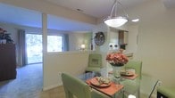 This single-floor residence offers a welcoming and well-appointed interior designed for comfortable everyday living. The dining area serves as a focal point of the open-concept layout, featuring a sleek glass-top table surrounded by light green upholstered chairs, elegantly set with warm-toned place settings and a floral centerpiece. A modern pendant light fixture hangs overhead, casting warm, inviting illumination across the space.

A half-wall partition thoughtfully separates the dining area from the adjacent living space, creating defined zones while maintaining an open, airy feel throughout. The living area beyond benefits from generous natural light streaming through a large window dressed with soft, flowing curtains, with a sliding glass door providing additional brightness and outdoor access.

The kitchen is seamlessly integrated into the layout, visible from the dining area and featuring wood-toned cabinetry and ample counter space. A decorative wine glass rack and round wall accent add personality to the transitional space between rooms.

Throughout the residence, neutral wall tones and light flooring contribute to a clean, cohesive aesthetic. The overall ambiance is bright, functional, and warmly styled, making it an inviting space suited for both everyday living and casual entertaining.