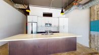This 2-bedroom, 2-bathroom urban loft delivers a compelling blend of industrial character and modern finishes across a single, thoughtfully designed floor. Polished concrete floors run throughout the space, complementing the exposed structural columns — some accented with bold color-blocked paint — and visible ductwork overhead that lend the unit an authentic warehouse aesthetic.

The kitchen serves as the centerpiece of the open-concept living area, featuring a large island with a rich, dark metal base and light stone countertop, white flat-front cabinetry, a classic subway tile backsplash, stainless steel appliances, and statement pendant lighting with gold interiors. A long, light-filled hallway connects the living areas to the private rooms, with industrial ceiling details adding visual interest throughout.

The bedrooms are generously proportioned, with one featuring large industrial-style multi-pane windows that flood the space with natural light, and another with a smaller clerestory window. Both bathrooms are finished in clean white palettes with subway tile — one includes a soaking tub with a glass-panel surround, while the other offers a walk-in shower with a bold black-framed glass enclosure and a wide double vanity. The overall ambiance is cool, urban, and contemporary.