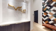The Villas Spa and Fitness Center is a thoughtfully designed amenity space that combines luxury wellness aesthetics with fully equipped athletic facilities. The spa reception area makes an immediate impression with its dramatic floor-to-ceiling accent wall clad in a geometric mosaic of multi-colored marble tiles in deep green, terracotta, white, and black. A sleek reception desk with dark fluted cabinetry and gold hardware sits beneath a white marble-paneled wall bearing the "Villas Spa" logo in brushed gold lettering. Light hardwood flooring and a cognac leather bench complete the refined entryway.

The expansive fitness center features soaring ceilings, large floor-to-ceiling windows that flood the space with natural light, and hexagonal pendant lighting. It is well-equipped with treadmills, ellipticals, cable machines, free weights, medicine balls, kettlebells, resistance equipment, and a punching bag. A textured wood-plank accent wall anchors the strength training zone, adding warmth to the industrial-modern aesthetic. A dedicated spin studio houses Peloton bikes, a large video display wall, and generous windows. Additional areas include a steam or sauna room with polished grey stone walls, rounding out a comprehensive wellness destination.