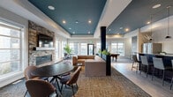 Located at Harbour Landing in Regina, Saskatchewan, this contemporary clubhouse is a thoughtfully designed communal space that balances comfort and functionality. The open-concept layout is anchored by a striking lounge area featuring a plush sectional sofa and armchairs arranged around a stone-clad linear fireplace, which serves as a focal point beneath a wall-mounted television. A signature tray ceiling finished in a deep teal hue with recessed pot lighting adds architectural depth and a refined sense of style to the main gathering area. Large windows line the perimeter, flooding the space with natural light and offering views of the surrounding neighbourhood. Patterned carpet tiles define the lounge zone, while light hardwood-style flooring transitions into the adjoining areas. A full-size pool table anchors a dedicated games area, complemented by a wood-panelled accent wall and bar-height seating. A kitchenette nearby features dark cabinetry, a stainless steel refrigerator, and pendant lighting, providing convenient amenities for entertaining. Structural columns with integrated planters add organic texture and subtly zone the space. Dining and café-style seating areas provide flexible options for social gatherings, making this clubhouse a well-equipped and inviting retreat for residents.
