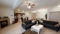 This single-floor space offers a thoughtfully designed open-concept layout that connects the living, dining, and kitchen areas into one cohesive, flowing environment. The living room serves as the central gathering space, featuring plush dark sofas, a clean white coffee table, and a ceiling fan with integrated lighting that keeps the area comfortable and well-lit. Neutral-toned carpeting adds warmth underfoot throughout the main living zones.

The kitchen is outfitted with rich dark cabinetry, white appliances including a range and refrigerator, and a raised breakfast bar with seating — a practical and inviting spot for casual meals. The kitchen flows naturally into the dining area, which is anchored by a rectangular table with upholstered seating and an elegant chandelier overhead, adding a touch of refinement to the space.

A door visible from the main living area hints at additional rooms or storage beyond the open floor plan. Recessed and ambient lighting throughout ensures the space remains bright and welcoming at all times. A decorative indoor tree adds a natural, lively accent to the dining corner. Overall, the layout is functional and well-proportioned, making excellent use of its single-level footprint for both everyday living and entertaining.