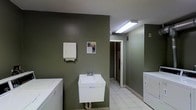This single-floor laundry facility is designed with practicality and functionality at its core. The space is finished with deep olive-green walls that lend a clean, utilitarian aesthetic throughout. Underfoot, light-colored ceramic tile flooring provides a durable and easy-to-maintain surface well-suited for high-use environments. Overhead fluorescent lighting ensures the area is well-lit, supporting comfortable use at any time of day. The room is equipped with multiple washing machines and dryers positioned along the walls, offering ample capacity for laundry needs. A wall-mounted utility sink with a standard faucet provides a convenient handwashing or pre-treatment station, accompanied by a paper towel dispenser mounted nearby for added hygiene. An open doorway leads to an adjoining area, suggesting additional utility or access space. A wall-mounted notice board displays informational materials, adding a community-oriented touch. The layout is straightforward and efficiently organized, maximizing usable floor space while keeping all essential amenities within easy reach. Ventilation is supported by a visible ceiling vent, ensuring air circulation within the room. Overall, this laundry facility is a well-equipped, no-frills space built to serve its users reliably and efficiently.