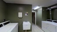 This single-floor laundry facility is designed with practicality and functionality at its core. The space is finished with deep olive-green walls that lend a clean, utilitarian aesthetic throughout. Underfoot, light-colored ceramic tile flooring provides a durable and easy-to-maintain surface well-suited for high-use environments. Overhead fluorescent lighting ensures the area is well-lit, supporting comfortable use at any time of day. The room is equipped with multiple washing machines and dryers positioned along the walls, offering ample capacity for laundry needs. A wall-mounted utility sink with a standard faucet provides a convenient handwashing or pre-treatment station, accompanied by a paper towel dispenser mounted nearby for added hygiene. An open doorway leads to an adjoining area, suggesting additional utility or access space. A wall-mounted notice board displays informational materials, adding a community-oriented touch. The layout is straightforward and efficiently organized, maximizing usable floor space while keeping all essential amenities within easy reach. Ventilation is supported by a visible ceiling vent, ensuring air circulation within the room. Overall, this laundry facility is a well-equipped, no-frills space built to serve its users reliably and efficiently.
