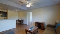This single-floor residence offers a practical and comfortable layout designed for everyday living. The living area serves as the central hub of the home, featuring warm hardwood-style flooring that flows throughout the space, lending a cohesive and inviting feel. A ceiling fan with an integrated light fixture is mounted overhead, providing both air circulation and ambient lighting to the room. The living space is open and connected to the dining area, which is positioned adjacent to a kitchen counter or breakfast bar, creating a seamless transition between cooking, dining, and relaxing. The dining area accommodates a wooden table and chairs, making it well-suited for shared meals. A media console provides additional storage and display space within the living area. A doorway visible from the main living space leads further into the home, suggesting additional rooms beyond. The walls are finished in a neutral, light tone that reflects light well and contributes to an airy, open ambiance. Overall, the space is straightforward in design, emphasizing functionality and ease of movement throughout the open-plan living and dining zones.