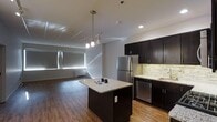 This contemporary single-floor space showcases a thoughtfully designed open-concept layout that seamlessly connects the kitchen and living areas. The kitchen is a standout feature, outfitted with rich dark cabinetry that contrasts elegantly against light granite countertops. A central island with matching granite provides additional workspace and a natural gathering point, illuminated by a stylish multi-pendant light fixture that hangs overhead. Stainless steel appliances, including a full-size refrigerator and dishwasher, lend a clean, modern finish. A textured stone tile backsplash adds visual depth and character to the cooking area. Recessed lighting throughout ensures the space is well-lit, complementing the warm hardwood flooring that runs across the kitchen and into the open living area. The living space is generously proportioned, with light-toned walls and large windows fitted with roller shades that allow for controlled natural light. The overall ambiance is sleek and modern, balancing functional design with contemporary aesthetics. The consistent use of dark and light tones throughout creates a cohesive, polished look that makes the space both inviting and visually striking.