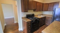 This single-floor space showcases a functional and well-designed kitchen as a central feature. The kitchen is outfitted with rich, dark-stained wood cabinetry that extends to the upper walls, providing generous storage space. The cabinetry is complemented by granite-style countertops in warm earth tones, adding a touch of elegance to the space. A black gas range with multiple burners serves as the kitchen's focal point, paired with a stainless steel sink set neatly into the countertop. The hardware throughout — drawer pulls and cabinet knobs — maintains a consistent brushed metal finish that ties the design together cohesively. The flooring consists of dark hardwood, which runs throughout the visible areas of the space, contributing to a warm and unified aesthetic. The neutral, light-toned walls provide a clean contrast against the darker cabinetry and flooring. A doorway visible in the background hints at additional rooms beyond the kitchen, suggesting a connected and practical floor plan. Overall, the space presents a classic design with durable, quality materials suited for everyday living.