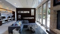 This elegant single-floor space exudes a refined, contemporary aesthetic from the moment you enter. Soaring ceilings amplify the sense of openness, while an expansive wall of floor-to-ceiling glass panels floods the interior with abundant natural light and offers unobstructed views of the surrounding outdoor environment. A dramatic dark wood feature wall serves as the room's focal point, housing illuminated display niches that showcase a curated collection of sculptural art pieces and decorative objects, adding depth and artistic character to the space. The seating area centers around a generous sectional sofa in a cool blue-grey tone, accented with an eclectic mix of throw pillows. Unique metallic tree-stump-style coffee tables anchor the seating arrangement atop a patterned area rug, blending organic forms with polished, industrial finishes. Recessed lighting complements the natural light, ensuring a well-lit ambiance throughout. The overall atmosphere is sophisticated and welcoming, striking a balance between modern luxury and approachable comfort, making this space well-suited as a high-end communal lounge or hospitality reception area.